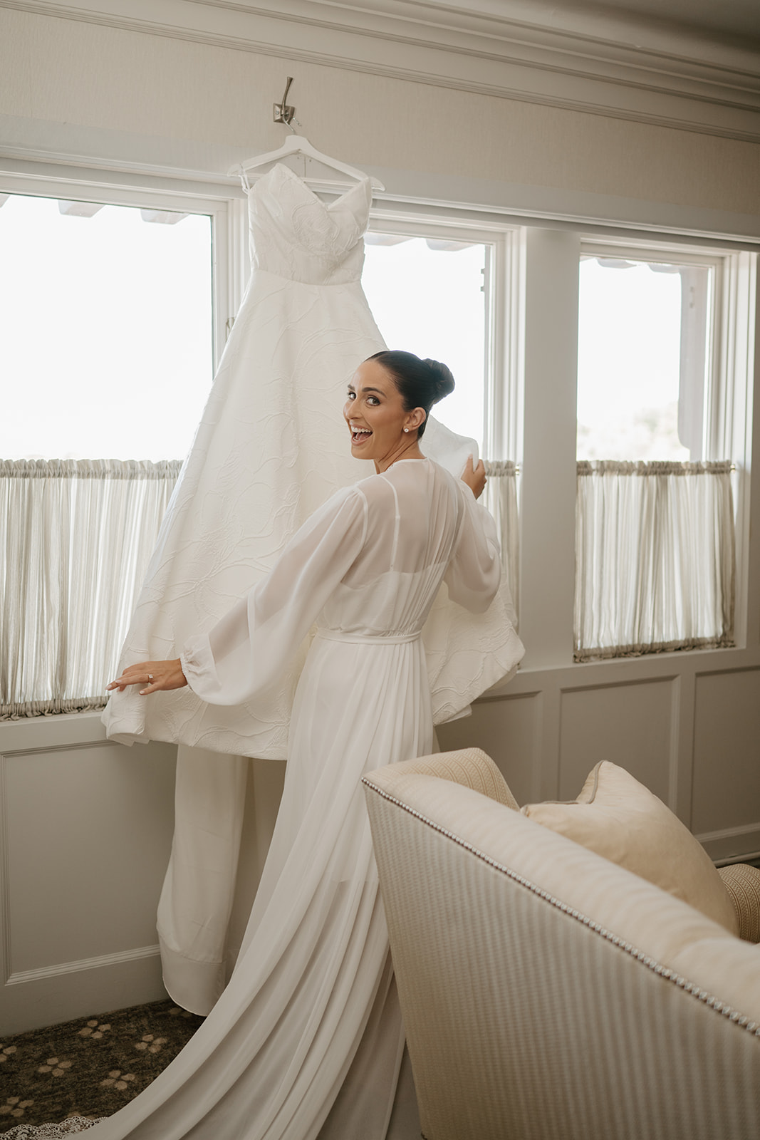 Excited bride in a white robe looking back while posing next to her hanging wedding gown.